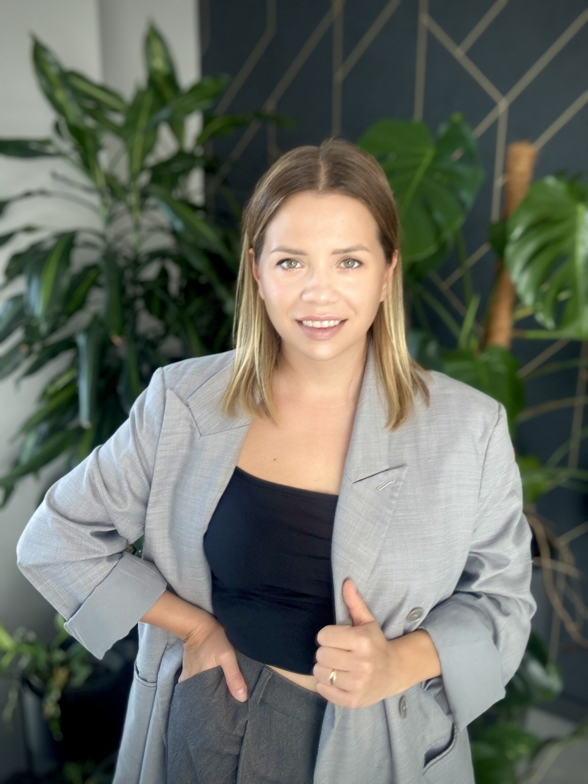 Justna Ciesielska - trenerka biznesu, stojąca na tle granatowo-złotej ściany i zielonymi roślinami