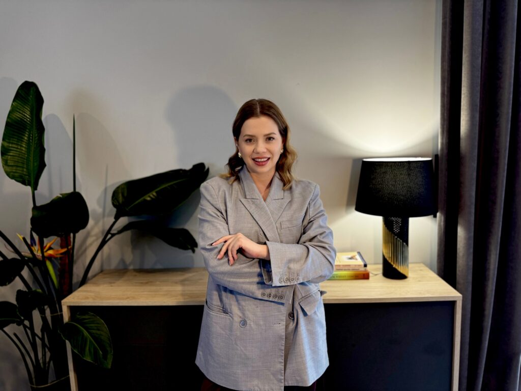 Justyna Ciesielska - trenerka biznesu, stojąca na tle biurka. Za nią delikatne oświetlenie i zielona roślina